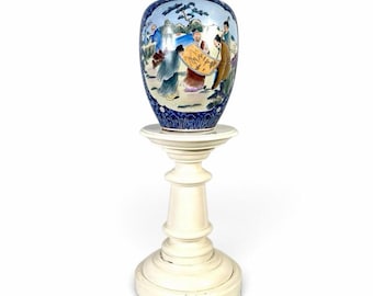May include: A porcelain ginger jar with a lid, featuring a hand-painted scene of figures in a landscape with blue and white floral patterns at the base. The jar rests on a cream-colored, pedestal-style wooden stand with a round base.