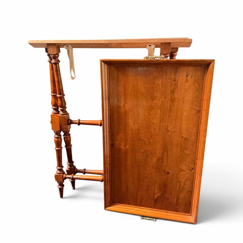 Puede incluir: Una mesa de bandeja de madera con soporte plegable. La bandeja rectangular tiene un acabado marr&oacute;n medio, un borde elevado y asas de metal dorado. El soporte cuenta con cuatro patas torneadas con refuerzos transversales.