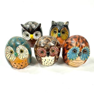 May include: A collection of five small, rounded owl figurines featuring hand-painted details. Each owl displays unique color patterns, including shades of blue, brown, orange, and black, with distinct facial markings and circular eye designs.