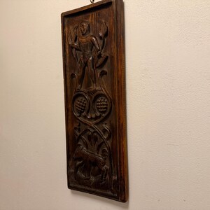 May include: A rectangular dark brown wooden wall plaque featuring a relief carving of a hooded figure holding a sickle, positioned above two circular floral motifs and a four-legged animal at the base, with a metal hanging loop at the top.