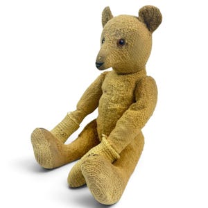 Pode incluir: Um urso de peluche de estilo vintage feito de tecido bege texturizado com um focinho longo, nariz costurado escuro e pequenas orelhas redondas. O urso apresenta punhos canelados desgastados nos bra&ccedil;os e pernas, olhos escuros e postura sentada.