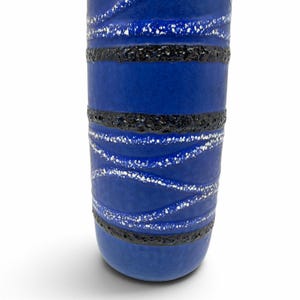 May include: A cylindrical ceramic vase featuring a vibrant blue base color. The surface is decorated with horizontal bands of textured black glaze and diagonal, wavy lines of speckled white glaze that wrap around the body of the vessel.