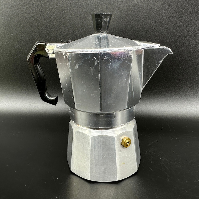 Puede incluir: Una cafetera de aluminio plateado con asa negra. La cafetera tiene una base redonda octogonal y un pico en el lateral.