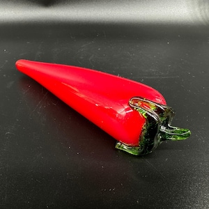 May include: A smooth, bright red glass chili pepper sculpture with a tapered, elongated body and a dark green glass stem attached at the wider end. The surface of the glass is glossy and reflective, showcasing a vibrant, solid red color throughout.