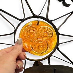 Peut inclure: Une applique murale en m&eacute;tal noir pr&eacute;sentant un motif en forme de soleil avec un insert en verre ambr&eacute; circulaire. Le centre en verre affiche trois motifs en spirale en relief. Le cadre en m&eacute;tal s'&eacute;tend en rayons fins et pointus.