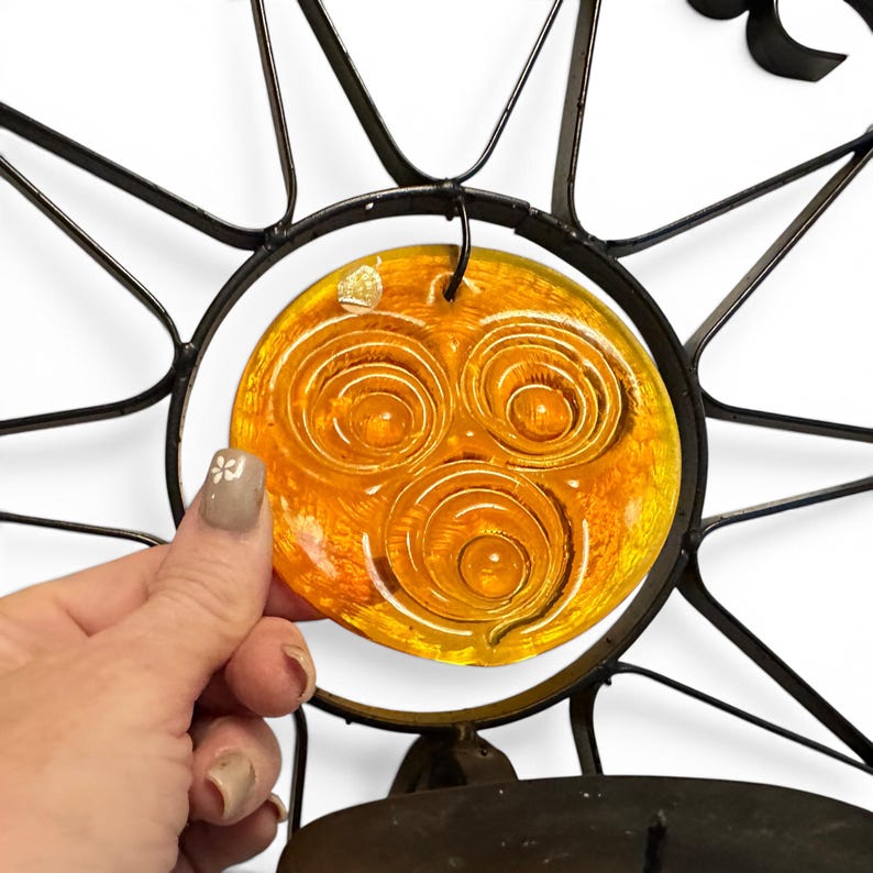 Peut inclure: Une applique murale en m&eacute;tal noir pr&eacute;sentant un motif en forme de soleil avec un insert en verre ambr&eacute; circulaire. Le centre en verre affiche trois motifs en spirale en relief. Le cadre en m&eacute;tal s'&eacute;tend en rayons fins et pointus.