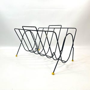 May include: Black metal magazine rack with a mid-century modern design. The rack features a series of curved wire supports and angled legs. The feet are tipped with small yellow caps. The rack is designed to hold magazines or other reading materials.