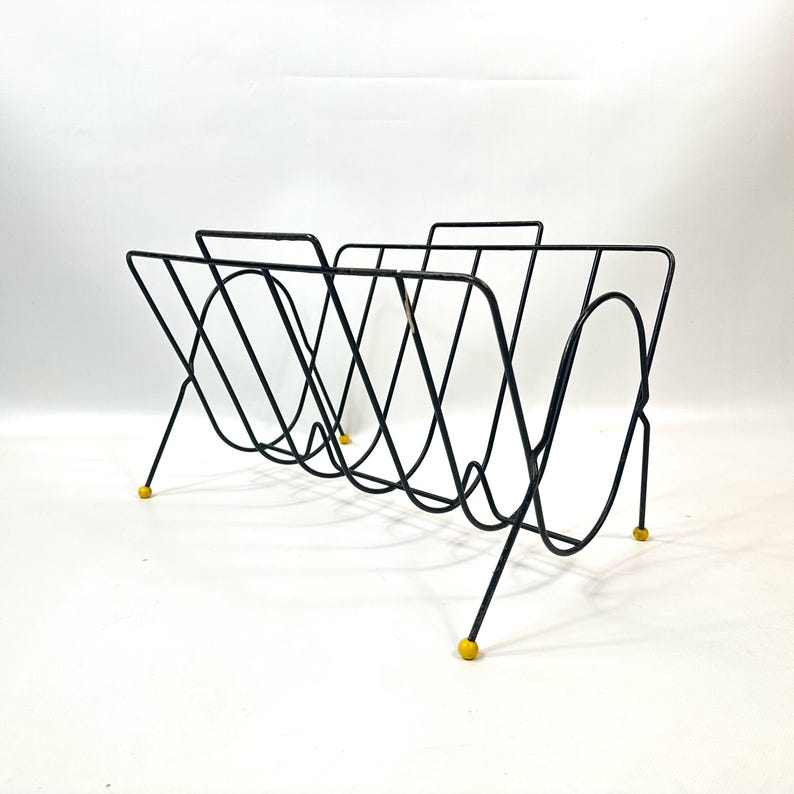 May include: Black metal magazine rack with a mid-century modern design. The rack features a series of curved wire supports and angled legs. The feet are tipped with small yellow caps. The rack is designed to hold magazines or other reading materials.