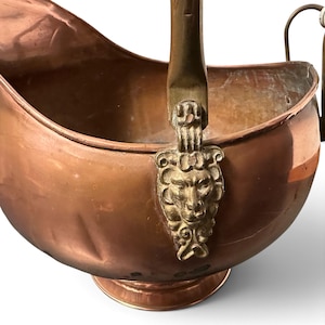 Pu&ograve; includere: Un secchio per carbone in rame con finitura lucida e corpo arrotondato. Il contenitore presenta un manico in ottone fissato ai lati con supporti decorativi in ottone a forma di teste di leone. Poggia su una base circolare con apertura ampia.