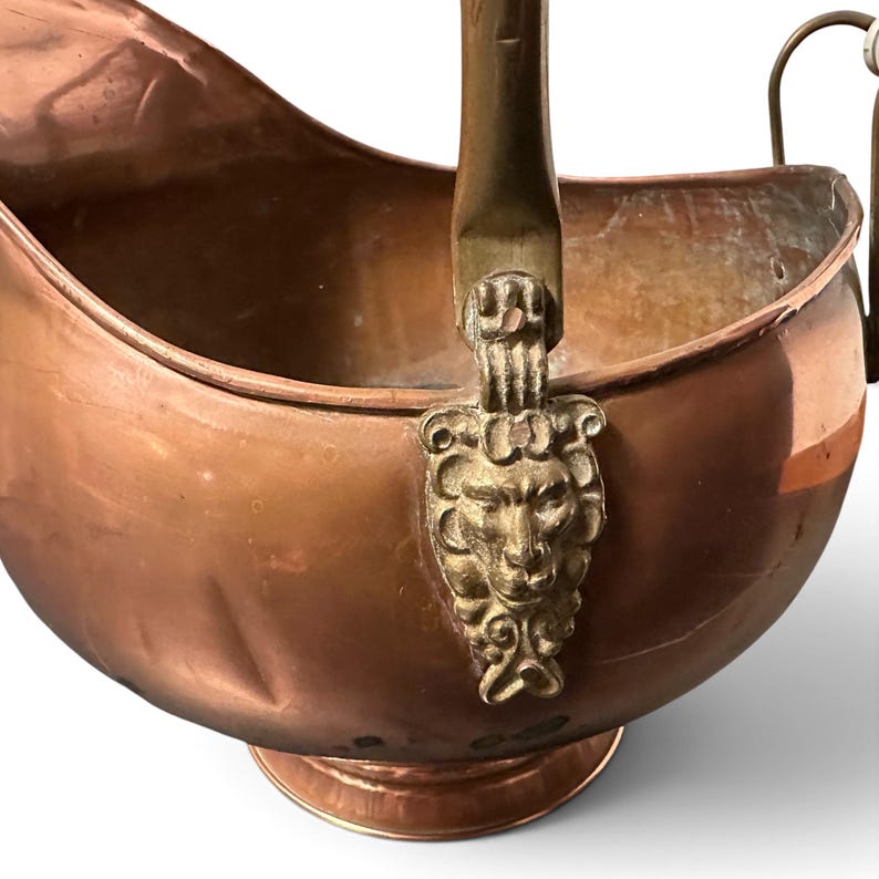 Pu&ograve; includere: Un secchio per carbone in rame con finitura lucida e corpo arrotondato. Il contenitore presenta un manico in ottone fissato ai lati con supporti decorativi in ottone a forma di teste di leone. Poggia su una base circolare con apertura ampia.