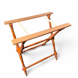 Puede incluir: Un soporte para equipaje de madera marr&oacute;n con un dise&ntilde;o plegable en forma de X, patas torneadas y dos rieles superiores horizontales conectados por dos correas de tela clara, ofreciendo una estructura firme para maletas.