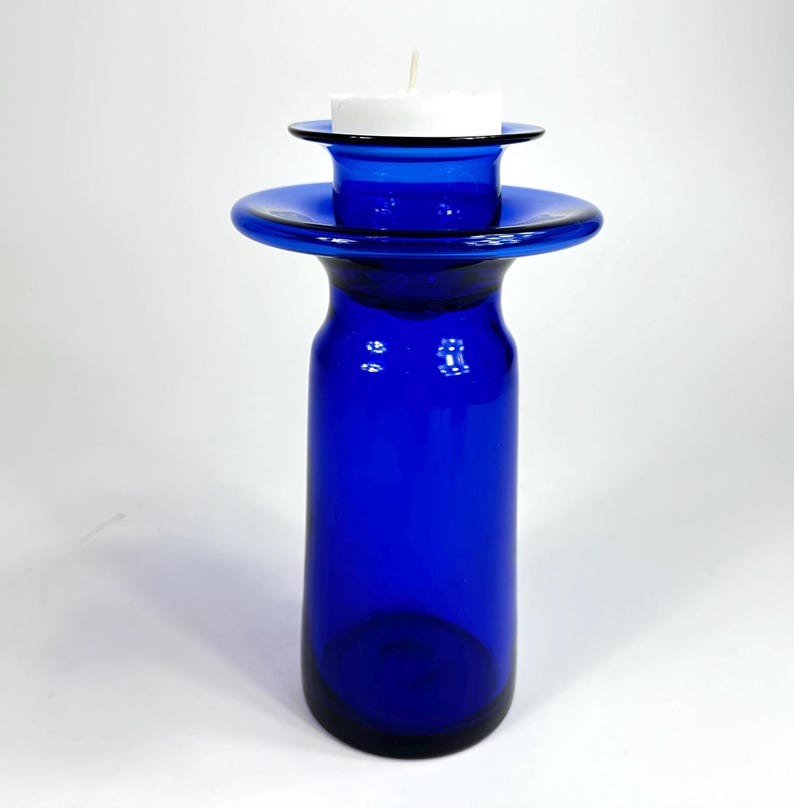 Op de afbeelding: Een kobaltblauwe glazen kaarsenhouder met een brede, platte bovenkant en een kleinere, ronde bovenkant om een kaars te houden. De kaarsenhouder is gemaakt van dik glas en heeft een gladde, gepolijste afwerking.