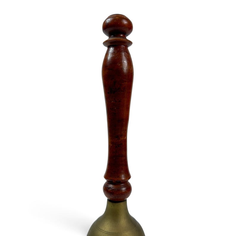 May include: A hand bell featuring a polished dark brown wooden handle with a rounded knob at the top and a flared base. The lower section consists of a metallic brass bell with a smooth, curved surface and a circular opening at the bottom.