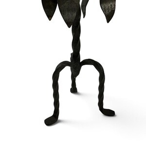 May include: A black wrought iron candle holder featuring a central twisted stem supported by three curved, twisted legs. The top section displays several pointed, leaf-shaped metal elements arranged in a circular pattern around the center.