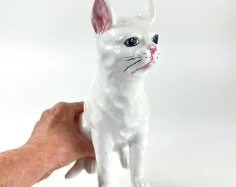 May include: A white ceramic cat figurine with pink accents on the ears and nose. The cat has black eyes and whiskers, and is posed in a seated position. The figurine is held by a person's hand.
