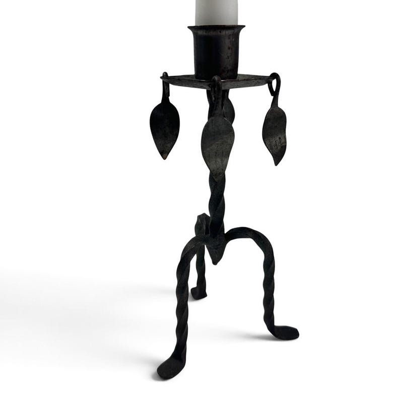 May include: A black metal candlestick holder featuring a central twisted stem, three curved legs, and a square top plate. The design includes hanging leaf-shaped metal accents suspended from the corners of the plate, supporting a single white candle.