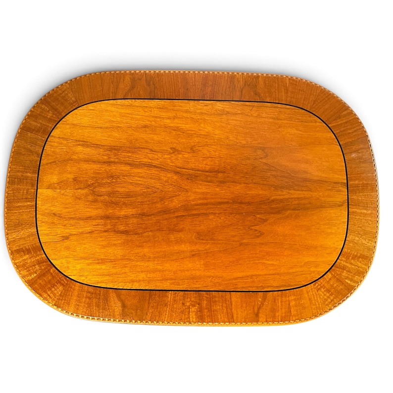 Op de afbeelding: Een ovaal houten dienblad met een glad, lichtbruin middenpaneel met een zichtbaar houtnerfpatroon. Het dienblad is omgeven door een brede, donkerbruine rand met een gestructureerd, herhalend patroon langs de buitenrand.
