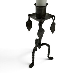 May include: A dark gray metal candle holder featuring a square top plate with a central cylindrical cup, a twisted central stem adorned with three metal leaf accents, and a tripod base composed of three twisted metal legs holding a white candle.
