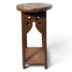 May include: A brown wooden plant stand featuring a circular top, four vertical legs, and a lower square shelf. The upper section of the stand includes decorative cutouts with circular holes and an arched opening between the legs.