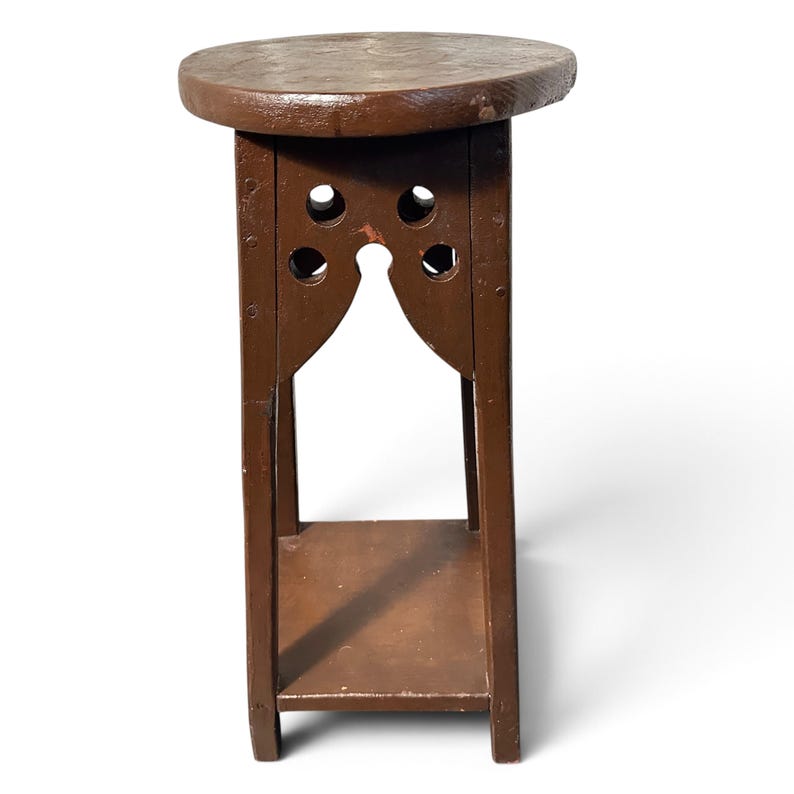 May include: A brown wooden plant stand featuring a circular top, four vertical legs, and a lower square shelf. The upper section of the stand includes decorative cutouts with circular holes and an arched opening between the legs.
