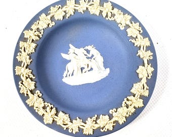 May include: A round blue ceramic plate featuring a raised white relief design. The center depicts a classical scene with figures and a horse, while the border is decorated with a continuous pattern of white grape leaves and vines in relief.