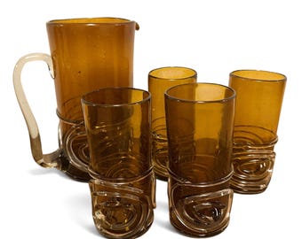 May include: A set of amber-colored glassware consisting of one pitcher and four tall glasses. Each piece features a clear glass handle or decorative base detail with a swirling, coiled design. The glass has a smooth, translucent finish.