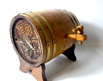 May include: A small wooden barrel with a horizontal orientation, featuring two metallic bands and a wooden spigot on the side. The front face displays a carved relief of a grape cluster with leaves, and the barrel rests on a wooden stand.