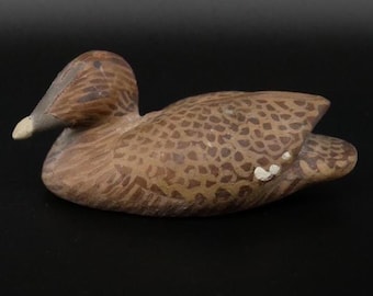 May include: A carved wooden duck decoy featuring a brown and tan mottled feather pattern across its body. The duck has a dark brown head, a light-colored beak tip, and a small white accent mark on the wing area, presented in a side profile view.