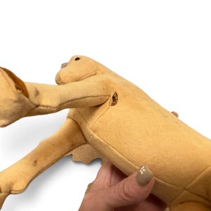 May include: A tan-colored plush animal toy with a smooth, felt-like texture. The figure features a long neck, a small dark eye, and brown accents on the limbs. There is a visible small tear or opening in the seam near the shoulder area.
