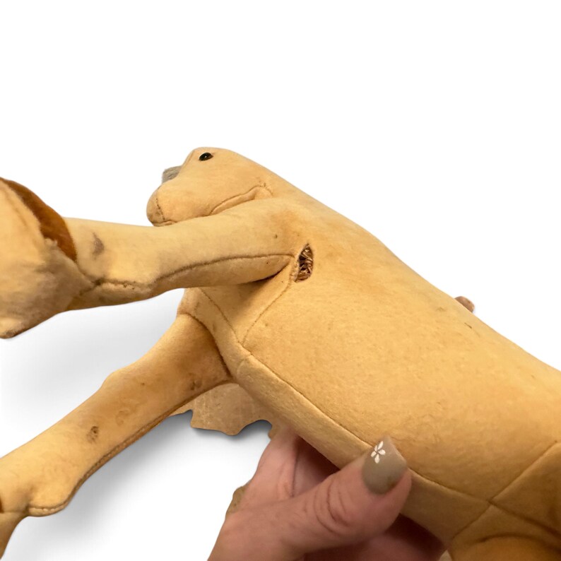 May include: A tan-colored plush animal toy with a smooth, felt-like texture. The figure features a long neck, a small dark eye, and brown accents on the limbs. There is a visible small tear or opening in the seam near the shoulder area.
