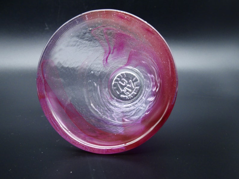 Puede incluir: Copa de vino de cristal transparente con un dise&ntilde;o en espiral de tonos rosas y rojos. La copa tiene un cuenco ancho y un dise&ntilde;o sin tallo. La parte inferior de la copa tiene una marca circular. La copa est&aacute; sobre un fondo oscuro.