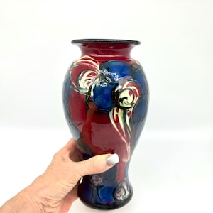 May include: A decorative ceramic vase with a glossy finish. The vase features a color palette of deep red, navy blue, and cream. The design includes swirling patterns and a black rim. The vase is approximately 10 inches tall.