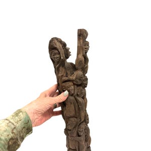May include: A tall, dark brown wooden sculpture featuring a vertical arrangement of multiple carved human figures stacked upon one another. The intricate carving displays various faces and bodies integrated into a single, textured wooden pillar structure.
