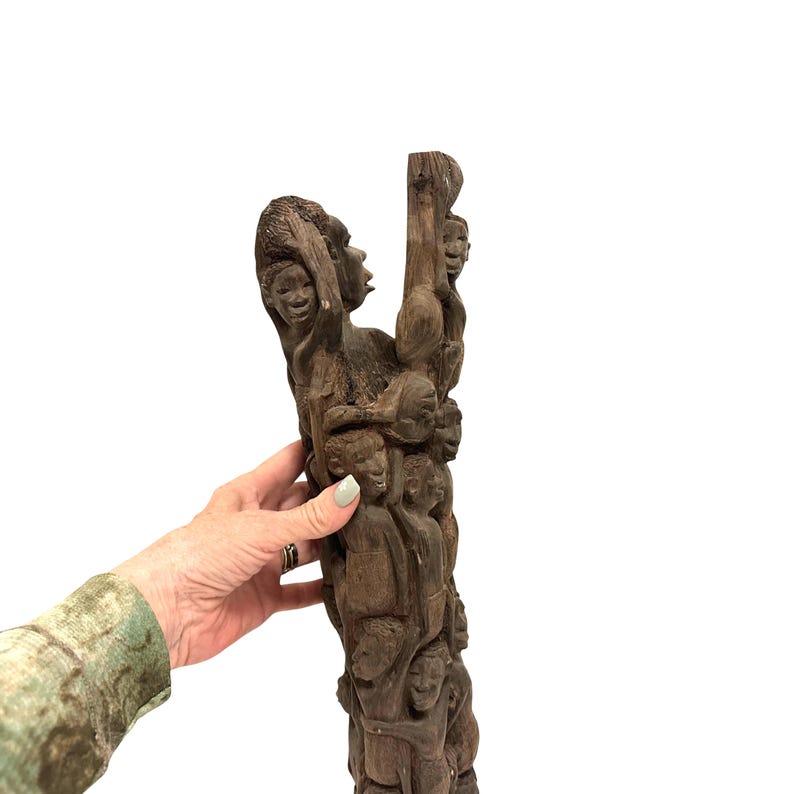 May include: A tall, dark brown wooden sculpture featuring a vertical arrangement of multiple carved human figures stacked upon one another. The intricate carving displays various faces and bodies integrated into a single, textured wooden pillar structure.