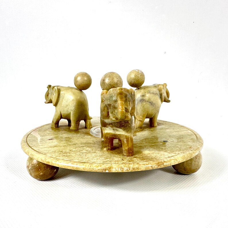 May include: A beige stone elephant figurine set. The set includes three elephant figurines and three spherical accents atop a circular base. The base is supported by four spherical feet. The elephants are carved with detailed features.