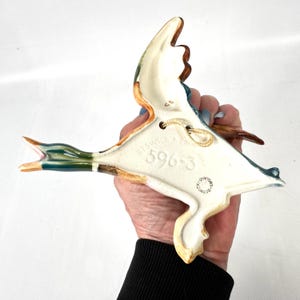 May include: A ceramic wall-mounted duck figurine in a flying pose with brown, green, and white glaze. The underside features the embossed text Beswick England 596-3 and a circular stamp reading Beswick England, with a beige hanging cord attached.