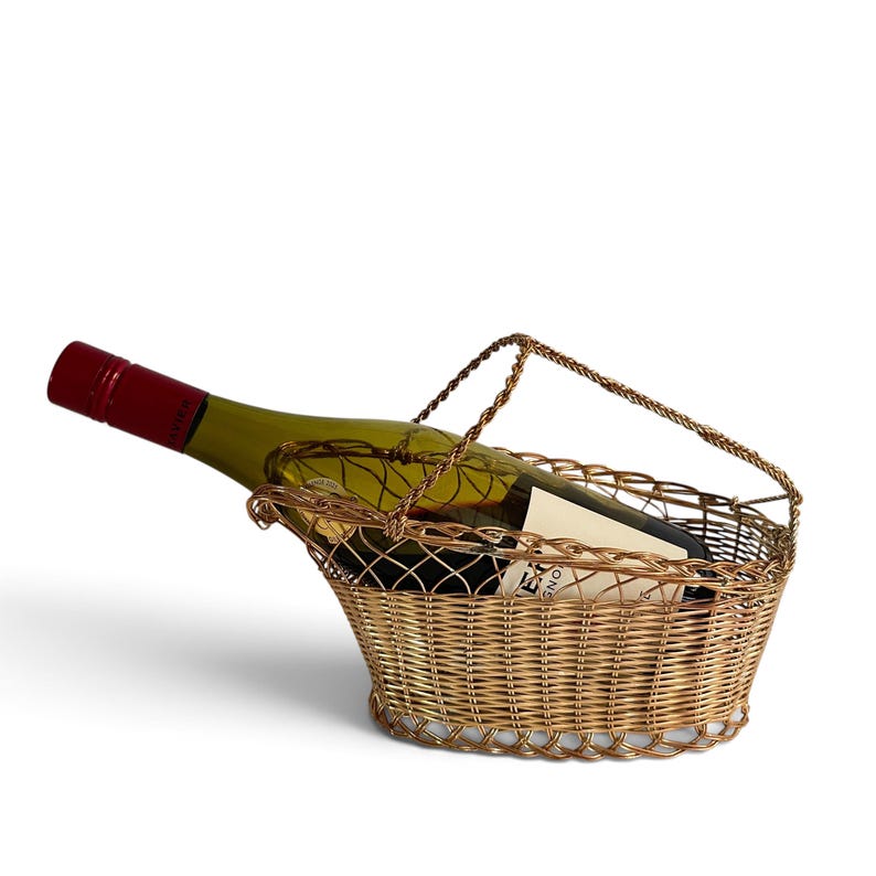 Puede incluir: Un soporte para botella de vino de alambre en tono dorado met&aacute;lico con un dise&ntilde;o de malla tejida y un asa arqueada. Una botella de vino de vidrio con tapa de rosca roja oscura y etiqueta color crema descansa horizontalmente en la cesta.