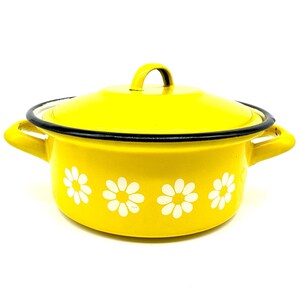 Pode incluir: Uma panela de esmalte amarelo brilhante com tampa a condizer e duas pegas laterais. O exterior apresenta um padrão repetitivo de flores de margarida brancas à volta da base. O rebordo da panela e a tampa têm um acabamento preto fino.