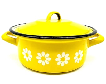 May include: A bright yellow enamel pot with a matching lid and two side handles. The exterior features a repeating pattern of white daisy flowers around the base. The rim of the pot and the lid are finished with a thin, contrasting black trim.