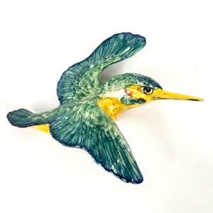 May include: A ceramic bird figurine in flight, featuring a detailed design. The bird has teal and green wings, a yellow breast, and a long yellow beak. The figurine is set against a white background, highlighting the vibrant colors and intricate details.