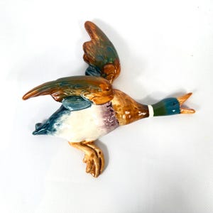 May include: A ceramic wall-mounted mallard duck figurine in flight, featuring a glossy finish with a dark green head, a white neck ring, brown and blue wings, and a white underbelly. The duck is depicted with its wings spread and beak open.