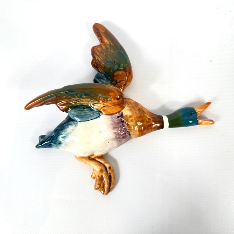 May include: A ceramic wall-mounted mallard duck figurine in flight, featuring a glossy finish with a dark green head, a white neck ring, brown and blue wings, and a white underbelly. The duck is depicted with its wings spread and beak open.