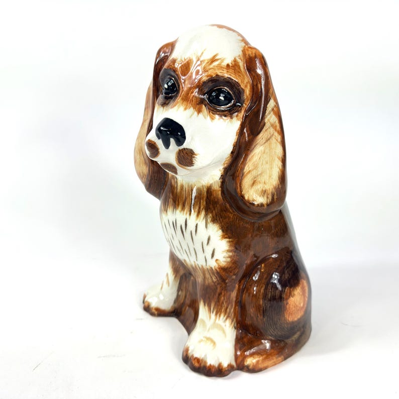 Peut inclure: Figurine en c&eacute;ramique repr&eacute;sentant un chien assis, avec des marques marron, blanches et beige. Le chien a de grands yeux expressifs et de longues oreilles tombantes. Finition brillante.