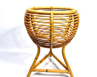 May include: A natural rattan plant stand with a round basket-like top and three curved legs. The stand is made of woven rattan and has a light brown color. The stand is designed to hold a plant pot.