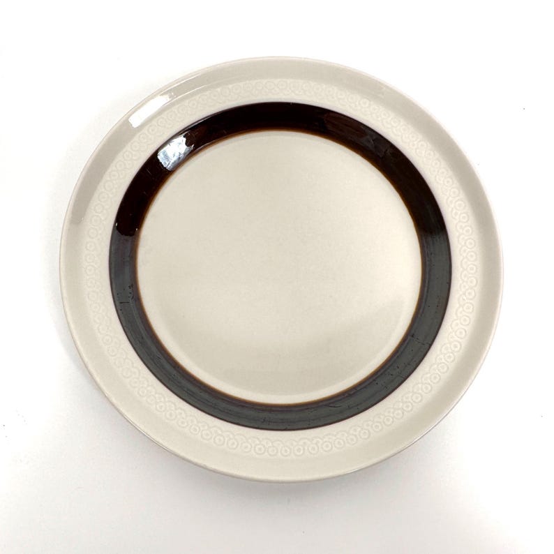 May include: A white ceramic plate with a brown band around the edge. The plate has a decorative pattern of small circles around the edge.