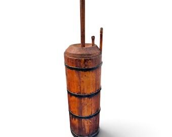 May include: A tall, cylindrical wooden butter churn featuring vertical staves held together by three dark metal bands. The churn includes a circular wooden lid with a central vertical plunger handle and a smaller secondary handle attached to the top.