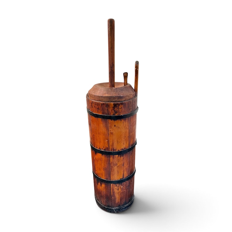 May include: A tall, cylindrical wooden butter churn featuring vertical staves held together by three dark metal bands. The churn includes a circular wooden lid with a central vertical plunger handle and a smaller secondary handle attached to the top.