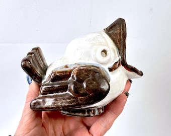 May include: A ceramic bird figurine with a white body and brown accents. The bird has an open beak and a hole on the side. The figurine is held in a hand.