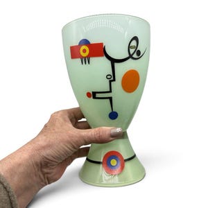 K&ouml;nnte beinhalten: Ein blass mintgr&uuml;ner Glaskelch mit abstrakten geometrischen Formen und Linien in Schwarz, Rot, Orange, Blau und Gelb. Das Design umfasst ein markantes rotes Rechteck mit einem gelben und blauen Kreis, einen gro&szlig;en orangefarbenen Kreis und ein kreisf&ouml;rmiges Emblem am Fu&szlig;.