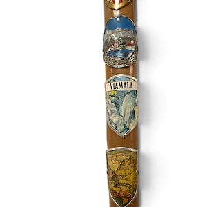 Puede incluir: Un bast&oacute;n de madera de color marr&oacute;n claro adornado con tres escudos met&aacute;licos de recuerdo. El escudo superior representa un paisaje monta&ntilde;oso, el central muestra un desfiladero con el texto VIAMALA y el inferior ilustra un paso de monta&ntilde;a.